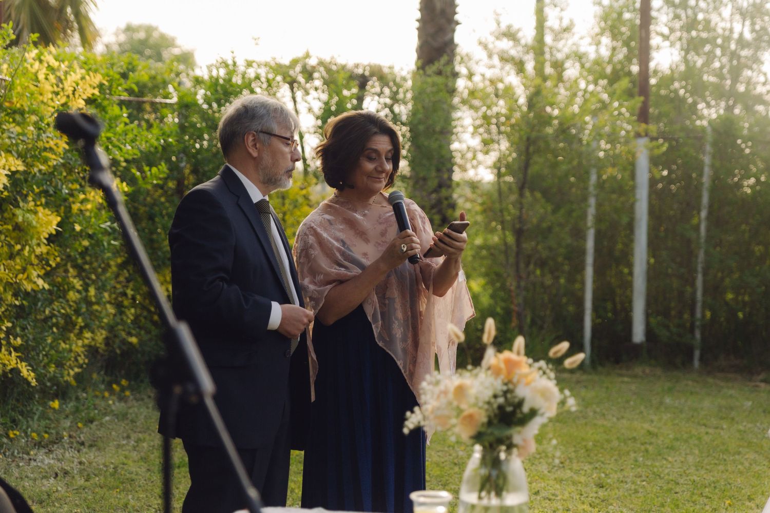 Older man in suit and woman in blue dress with shawl share a moment looking at a phone outdoors.