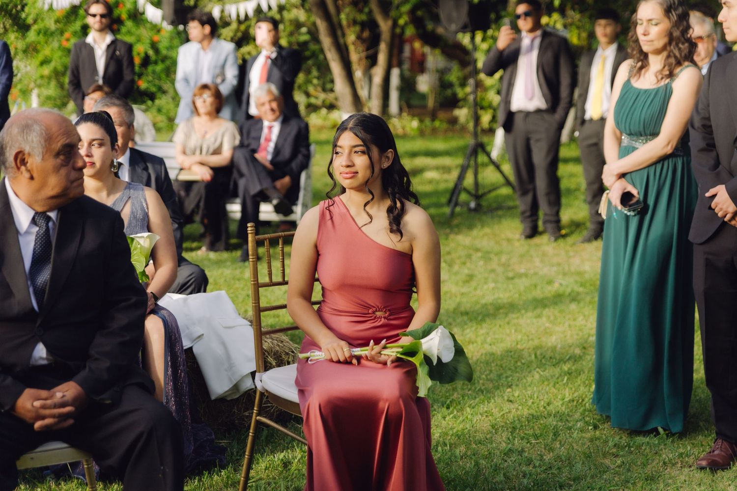 Wedding guests seated and standing outdoors, woman in red one-shoulder dress in foreground, green dress visible behind.