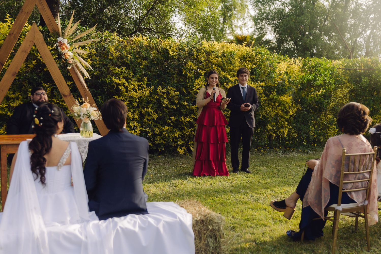 Woman in red dress speaks at outdoor garden wedding ceremony while bride and groom sit facing her on hay bales.