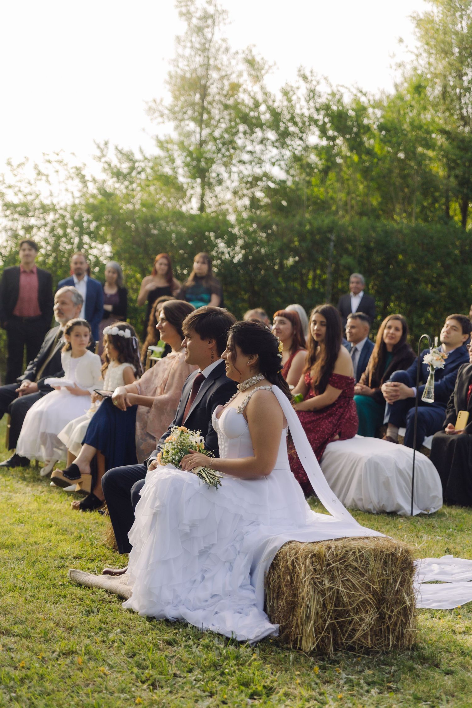Bride in white gown and groom sit on hay bales during outdoor rustic wedding ceremony with guests seated behind.