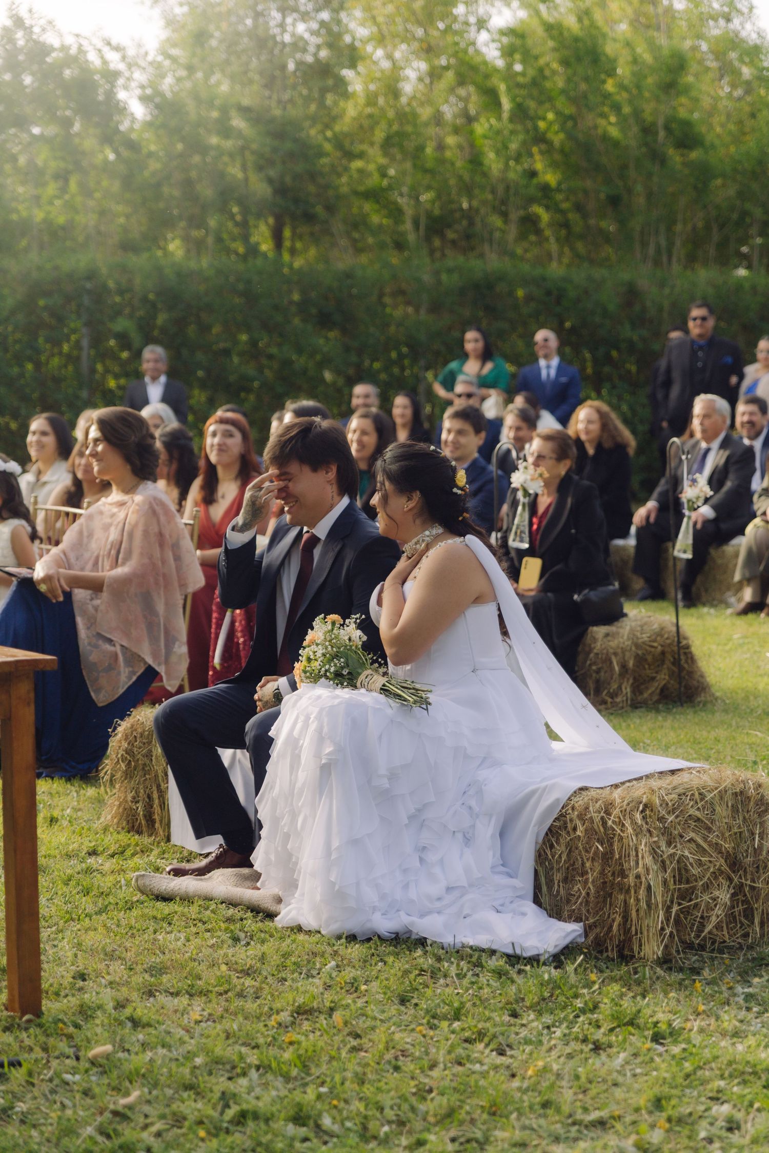 Bride holding bouquet and groom share a tender moment on hay bales during outdoor wedding ceremony with guests.