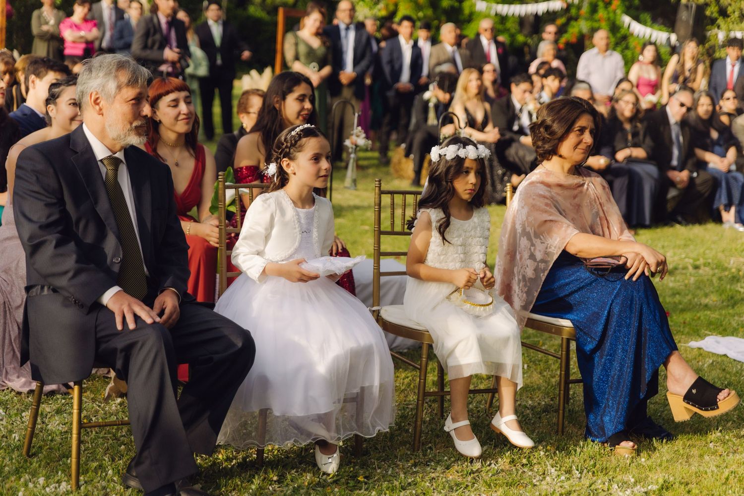 Two young flower girls in white dresses sit with adults during an outdoor garden wedding ceremony.