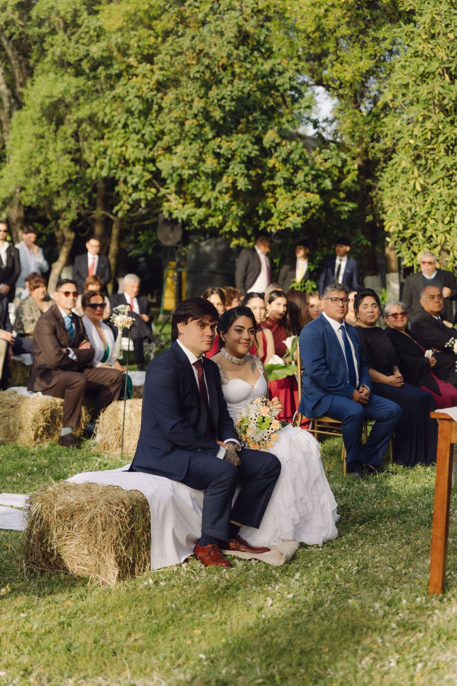 Bride and groom sit on hay bales during sunny outdoor rustic wedding ceremony surrounded by guests on grass.