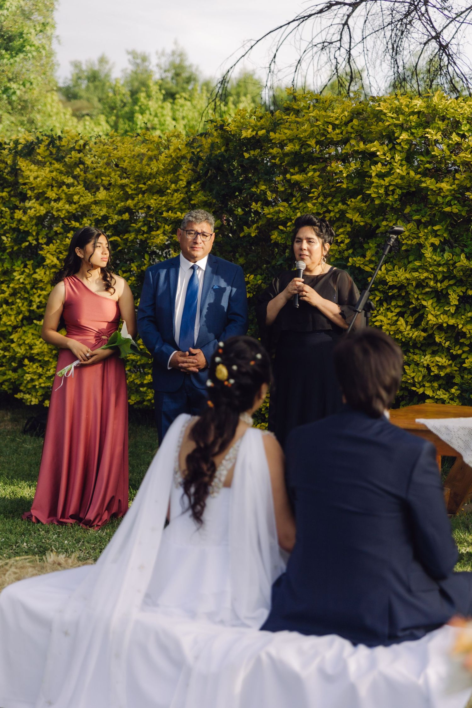Wedding officiant speaks into microphone flanked by attendants during outdoor garden ceremony with bride and groom.