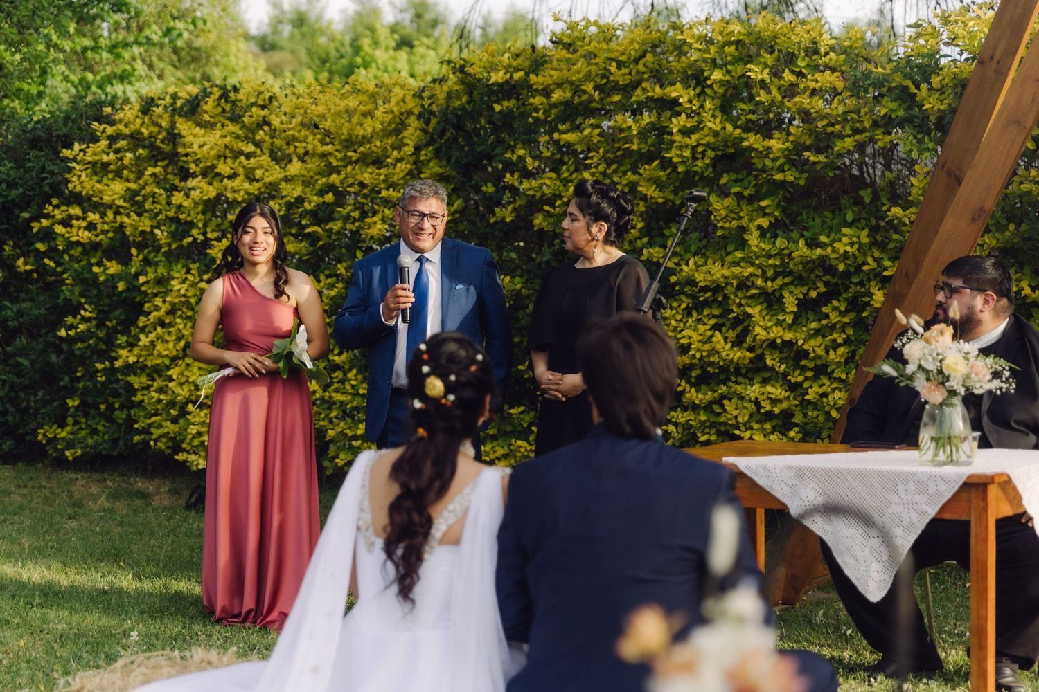 Wedding officiant addresses bride and groom during an outdoor garden ceremony with lush green hedges in background.