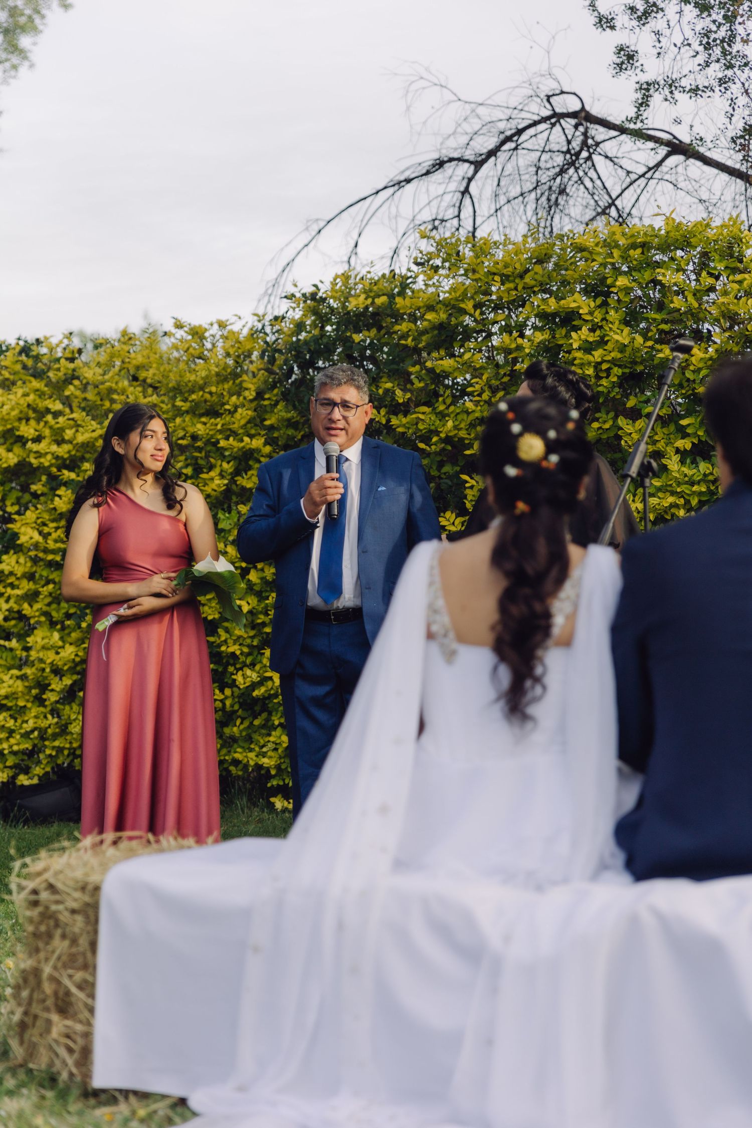 Man in blue suit speaks at outdoor wedding ceremony while woman in pink dress stands beside him at altar.