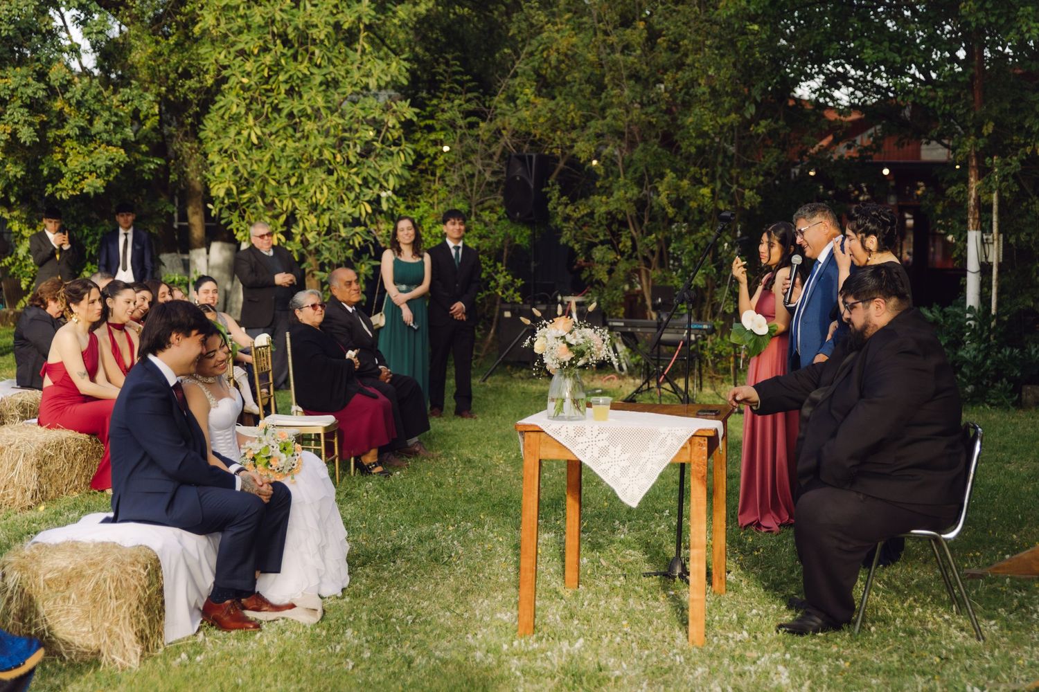 Wide shot of outdoor rustic wedding ceremony with guests seated on hay bales in a lush green garden setting.