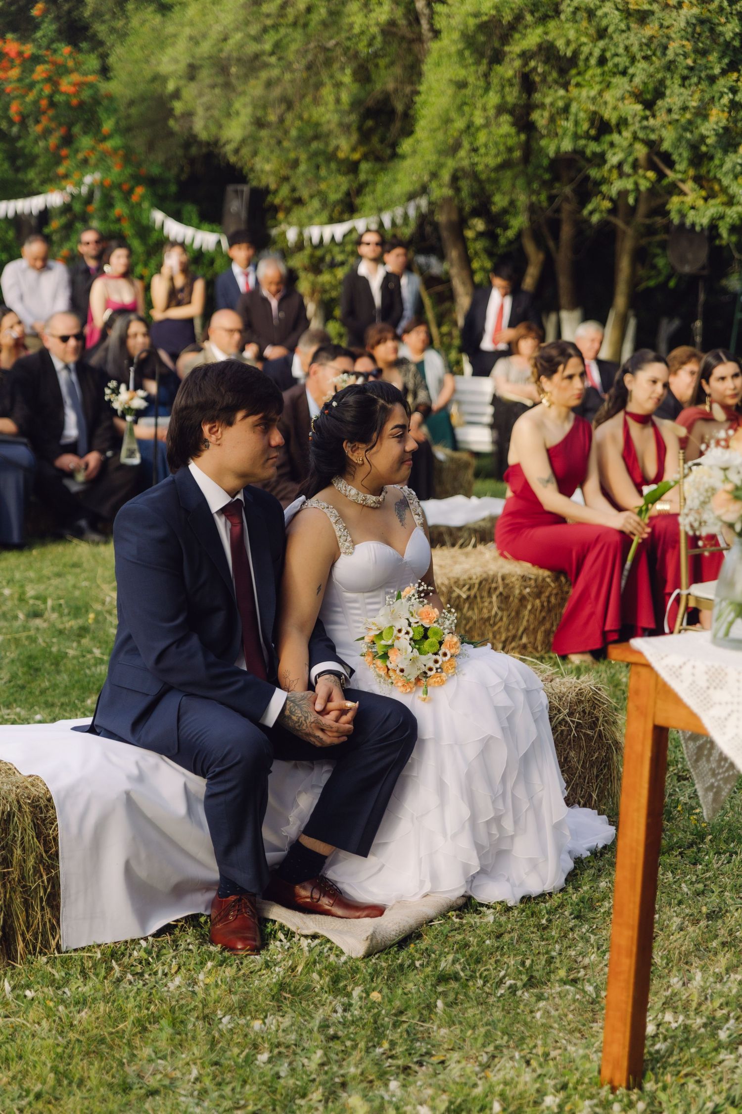 Bride with bouquet and groom sit closely on hay bales during outdoor wedding ceremony with guests and bridesmaid nearby.