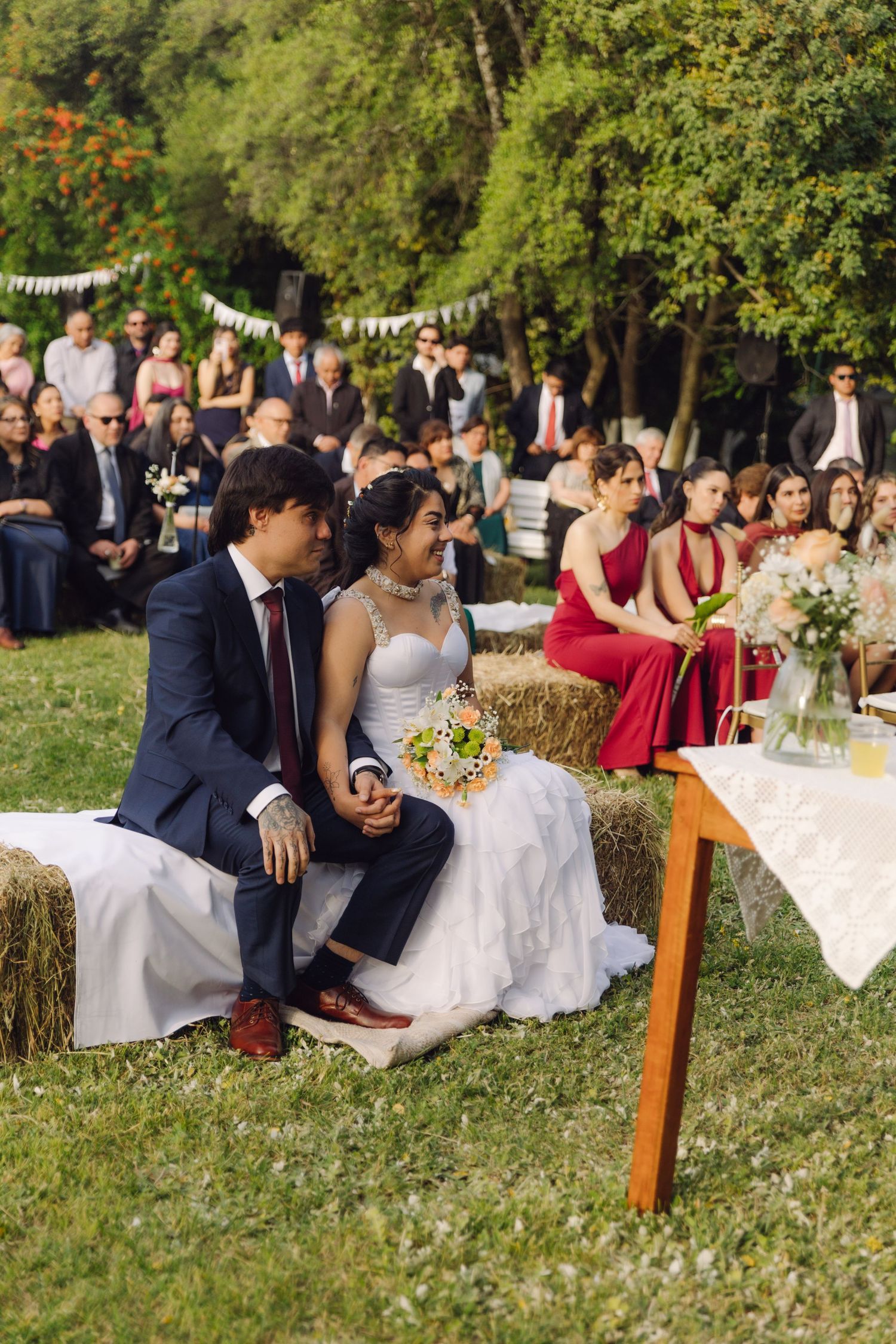 Bride and groom sit on hay bales during an outdoor rustic wedding ceremony with guests seated in background.