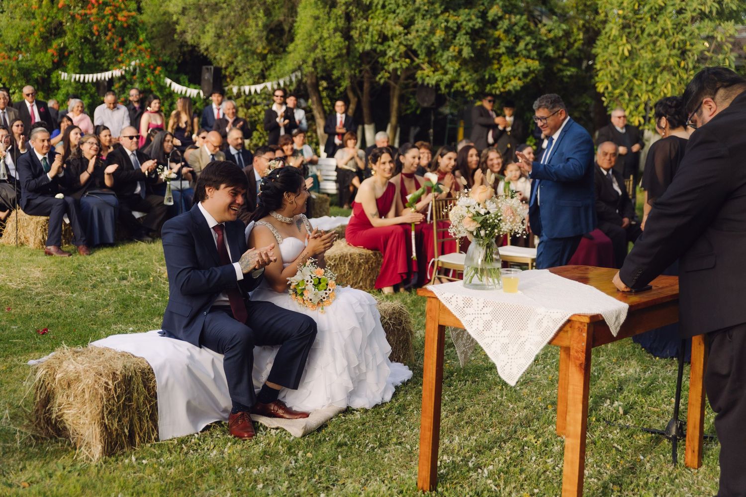Couple exchanges vows at outdoor rustic wedding ceremony seated on hay bales with officiant and guests present.