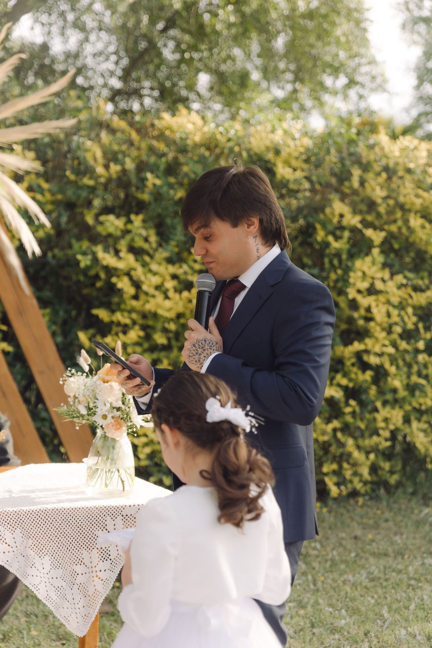 Wedding officiant reads vows into microphone while holding tablet at an outdoor garden ceremony.
