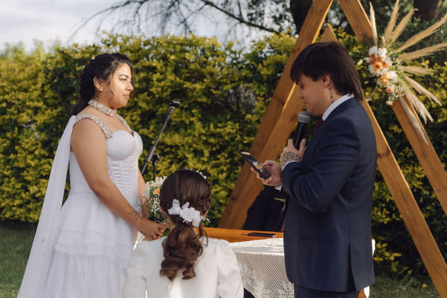 Bride and groom exchanging vows at outdoor wedding ceremony beneath wooden arch with greenery.