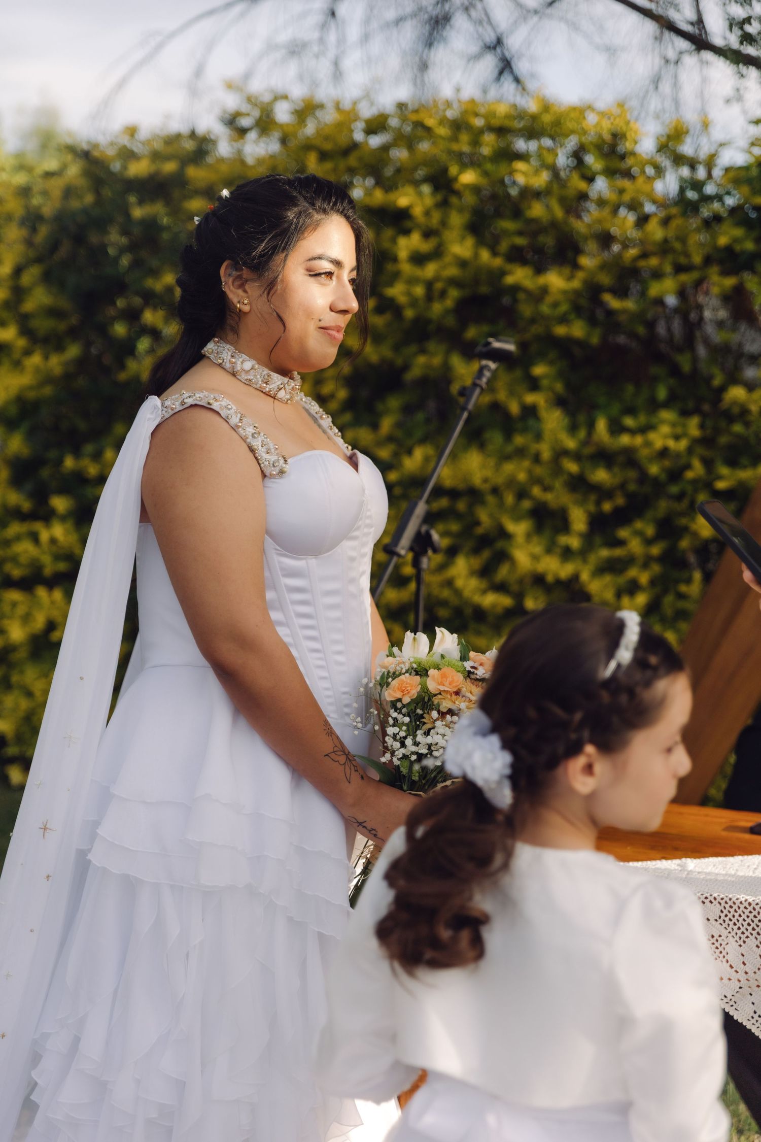 Bride in white gown holding peach bouquet stands outdoors at garden wedding ceremony with flower girl nearby.