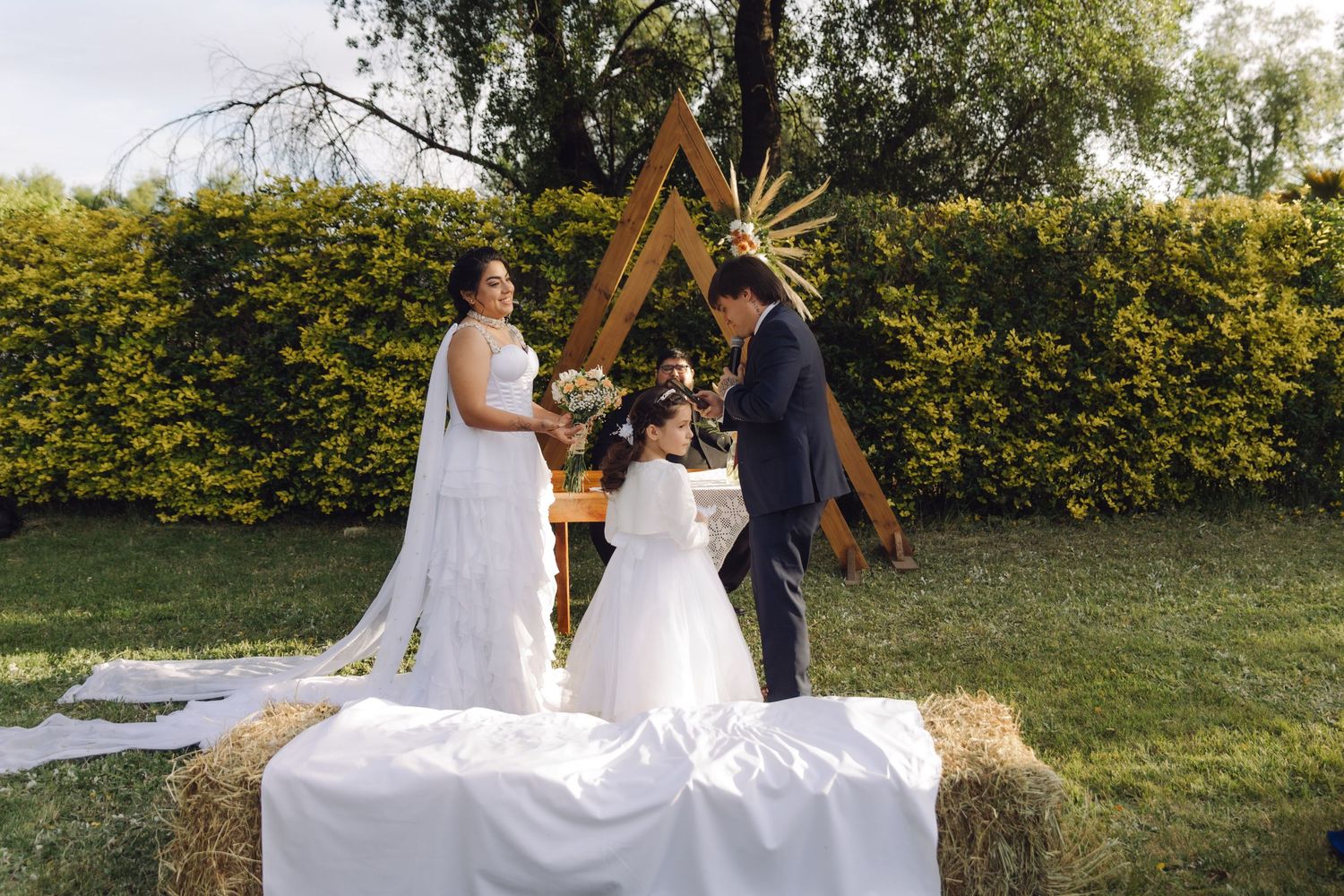 Bride and groom exchange vows with flower girl at rustic outdoor wedding altar on hay bales.