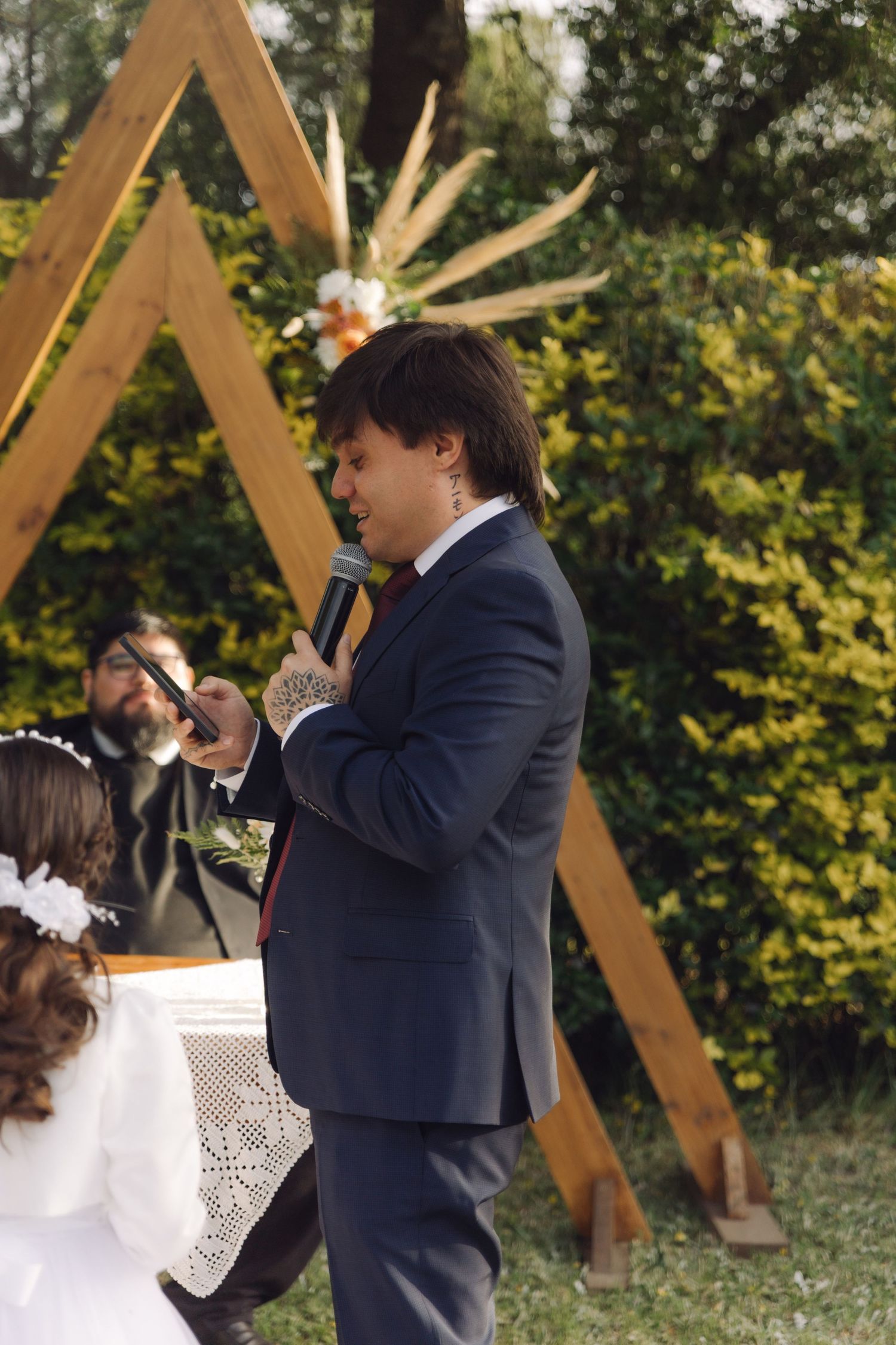 Groom in navy suit speaks into microphone at outdoor wedding ceremony before wooden triangular arch.