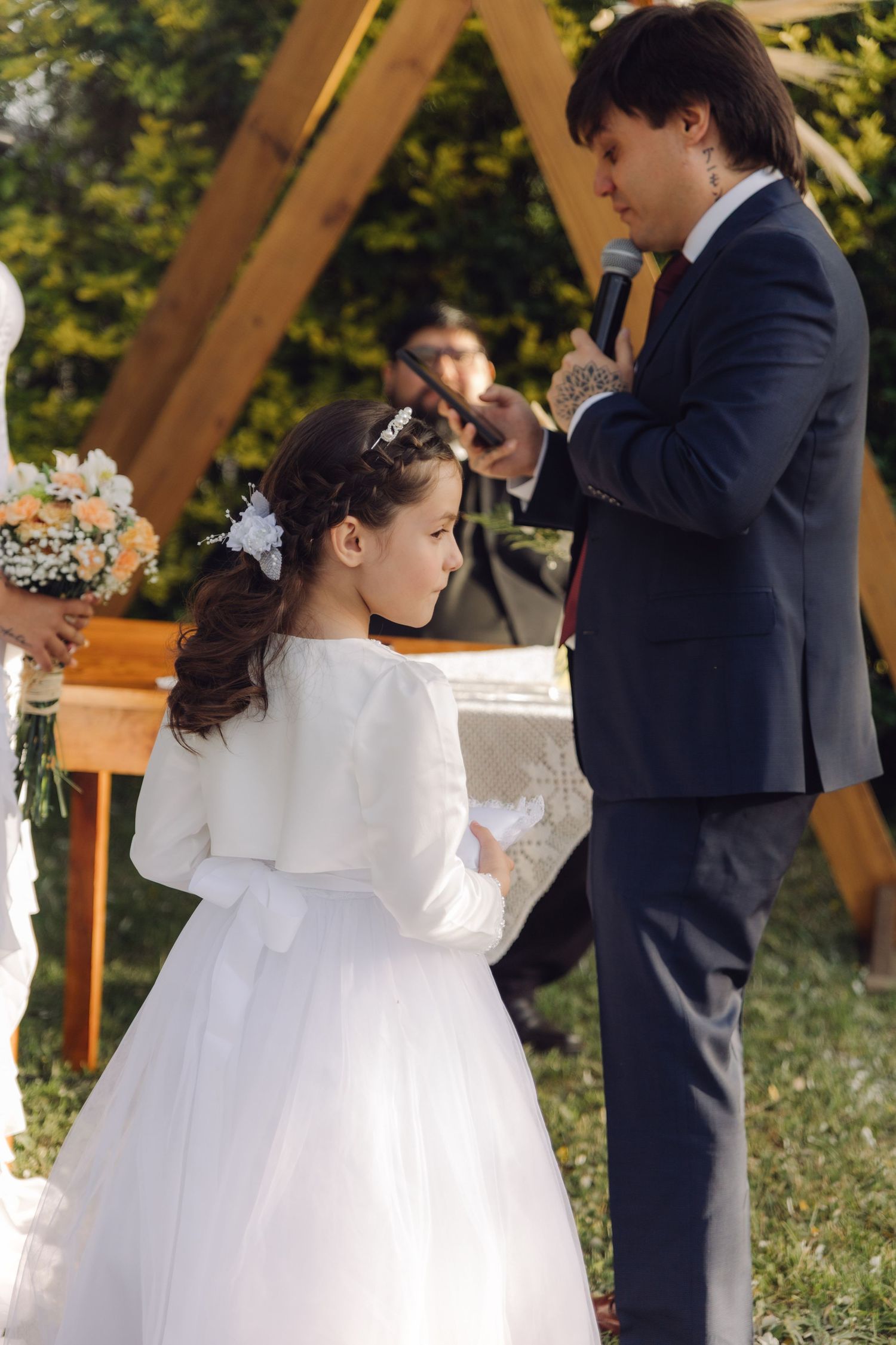 Young flower girl in white dress faces groom speaking at microphone during outdoor wedding ceremony.