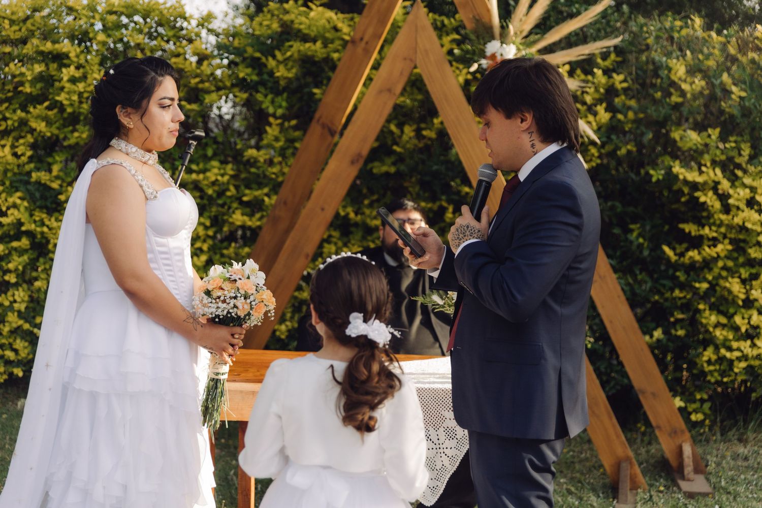Bride in white gown and groom in navy suit exchange rings with flower girl at outdoor ceremony.