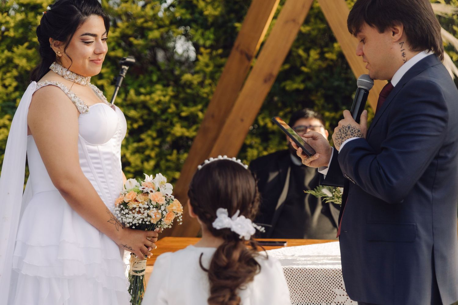 Groom reads vows to bride holding peach bouquet with flower girl between them at outdoor wedding.
