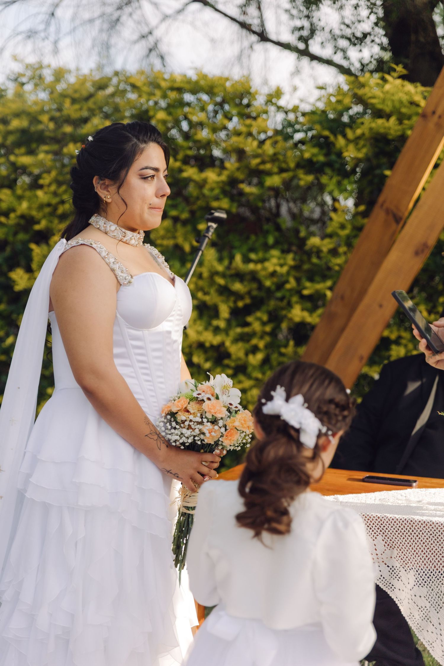 Elegant bride in white corset gown holds peach bouquet beside flower girl at outdoor garden ceremony.