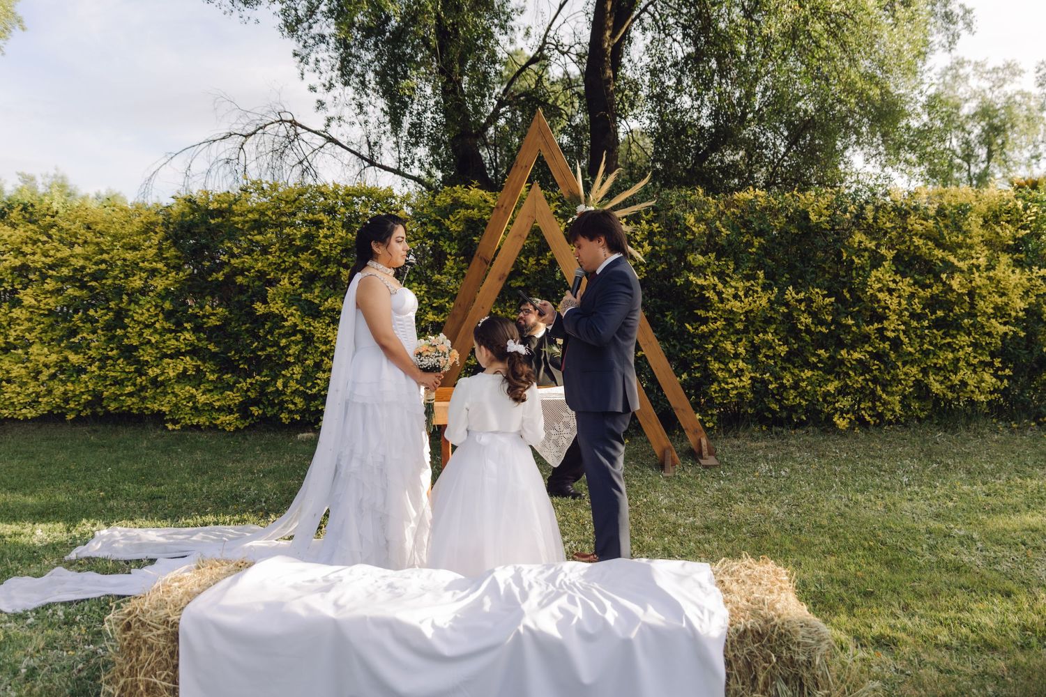 Bride groom and flower girl stand at rustic hay bale altar beneath wooden triangular arch outdoors.