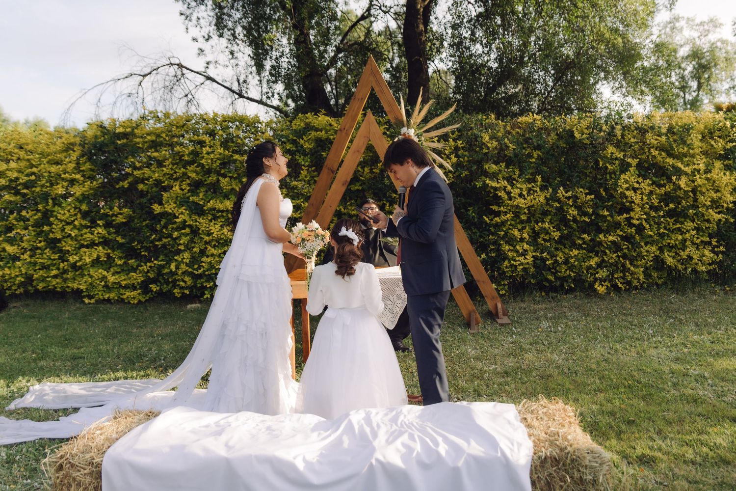 Bride groom and flower girl participate in outdoor wedding ceremony at wooden triangular arch altar.