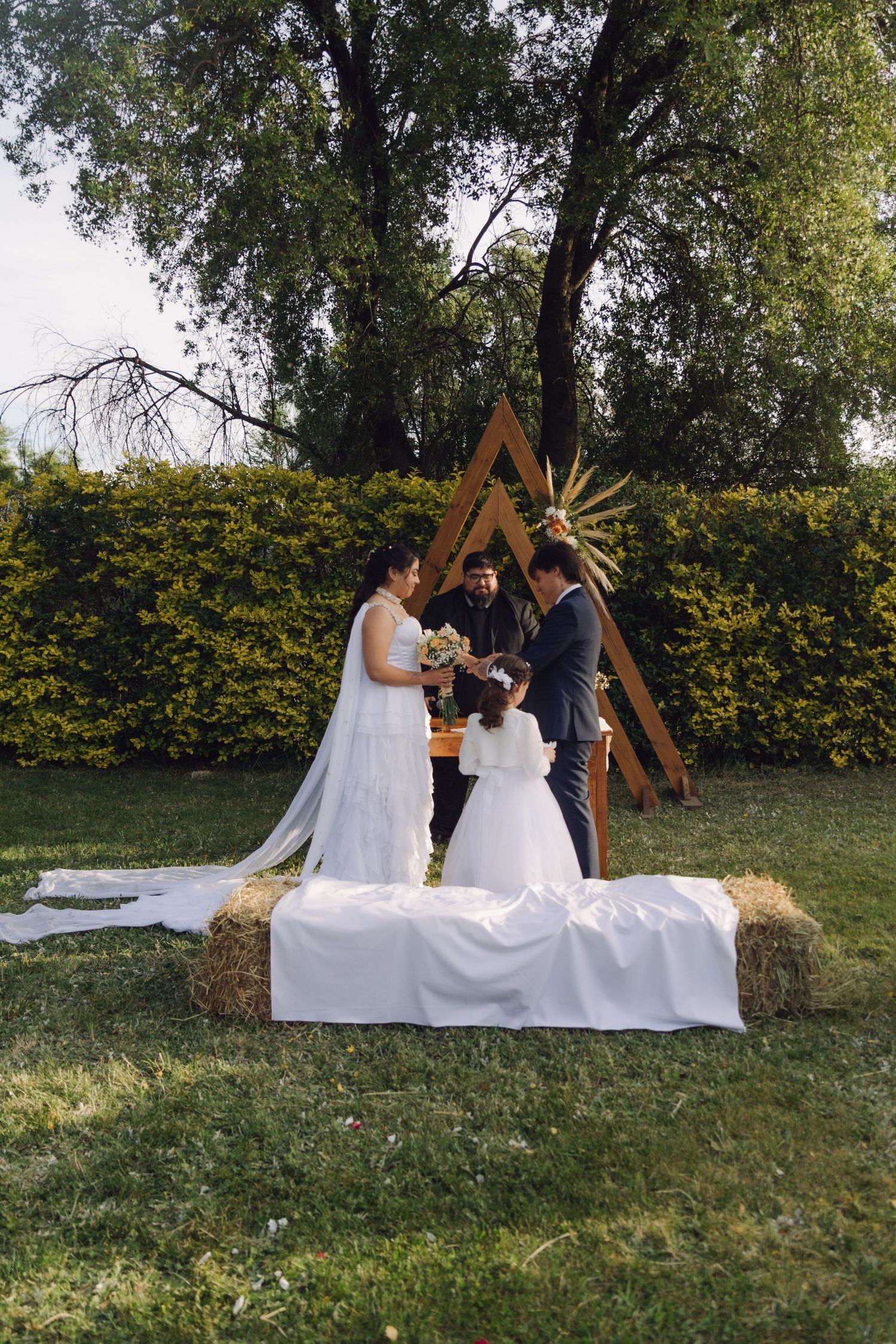 Bride and groom with officiant and flower girl at rustic outdoor wedding ceremony on hay bale altar.