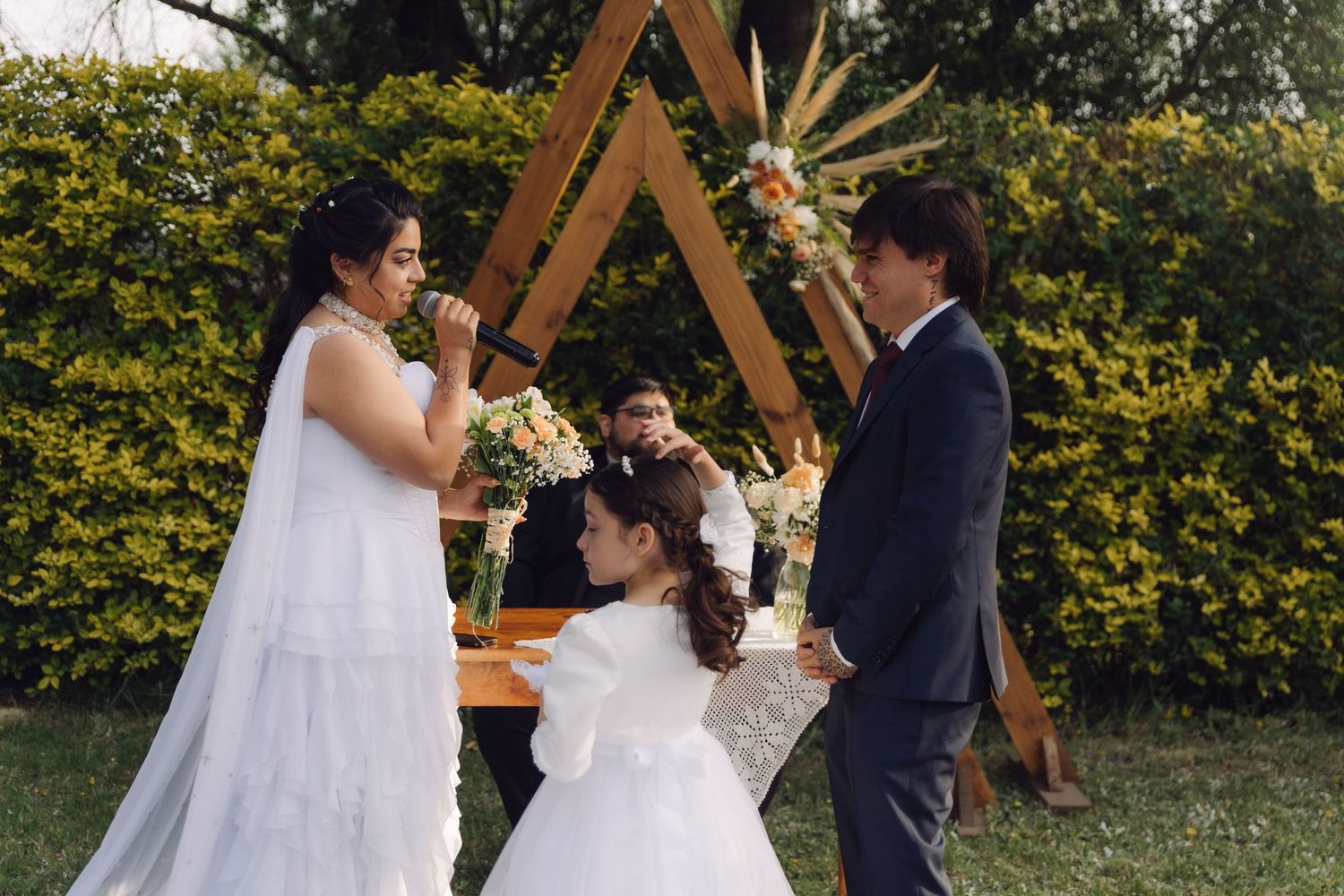Bride and groom exchange vows at outdoor wedding ceremony with triangular wooden arch backdrop and flower girl.