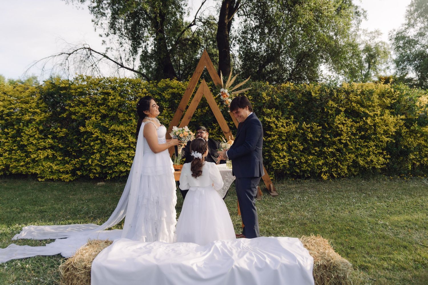 Couple stands at rustic outdoor wedding altar with triangular wooden arch on hay bales surrounded by greenery.