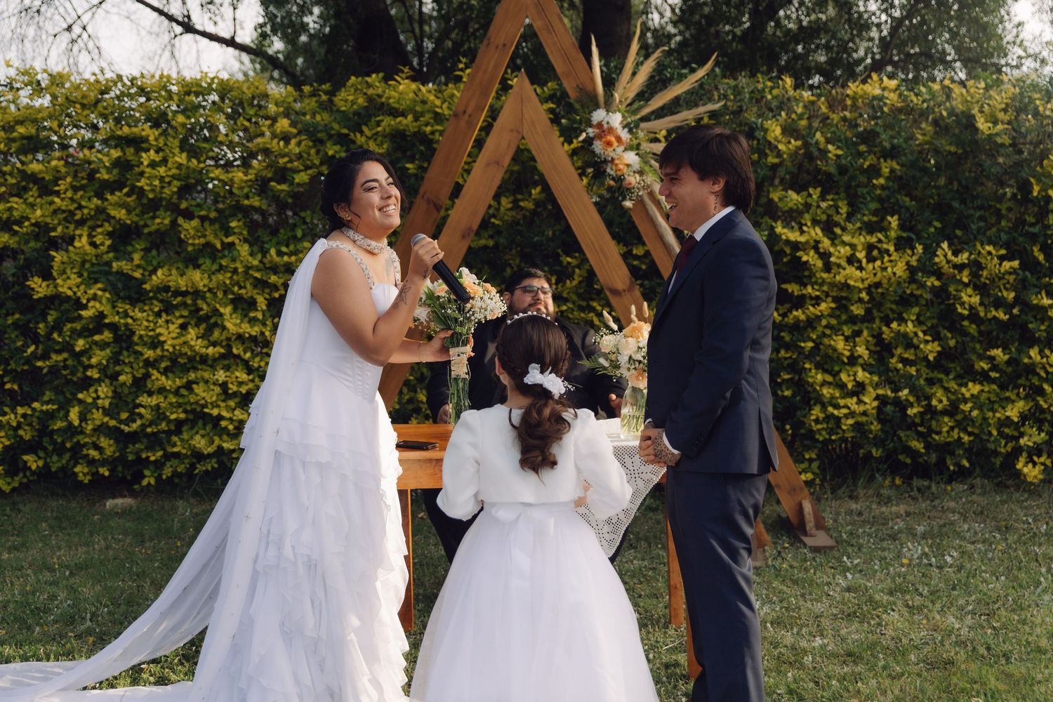 Bride in white tiered gown holds bouquet facing groom at boho outdoor wedding ceremony with wooden arch.