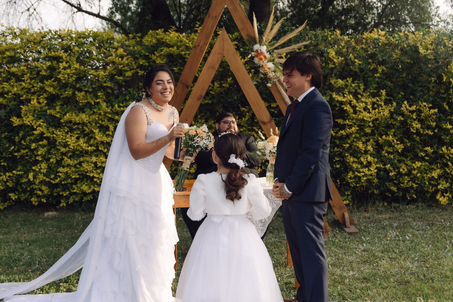 Smiling bride and groom share a joyful moment with flower girl during outdoor garden wedding ceremony.
