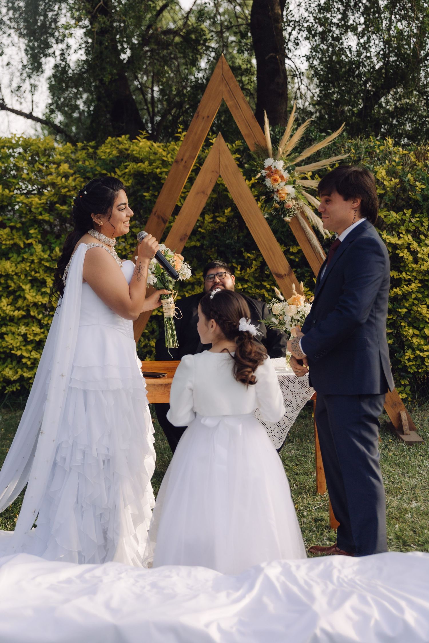 Bride and groom face each other at boho outdoor wedding with triangular wooden arch and dried floral decor.