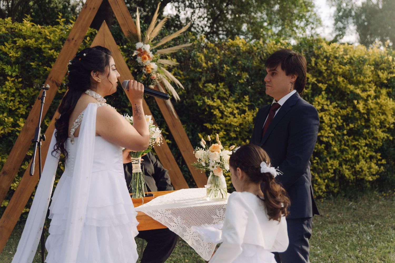 Bride reads vows at outdoor wedding ceremony beside triangular wooden arch with groom and flower girl present.