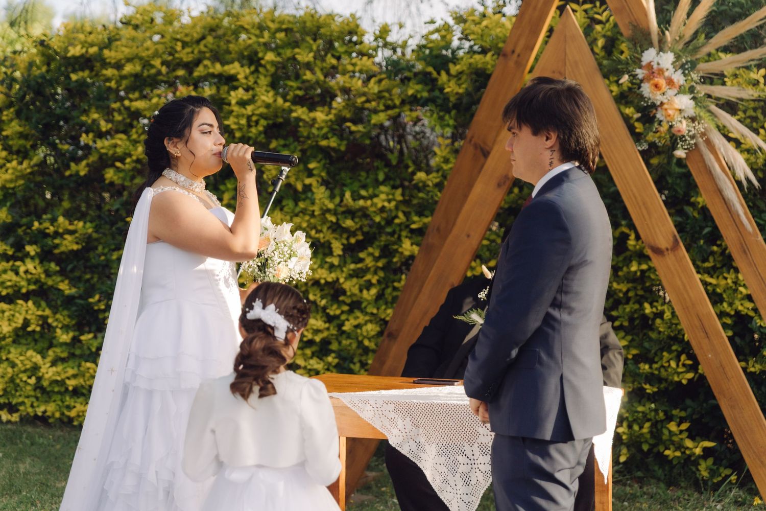 Emotional bride speaks vows into microphone facing groom at intimate outdoor boho wedding ceremony.