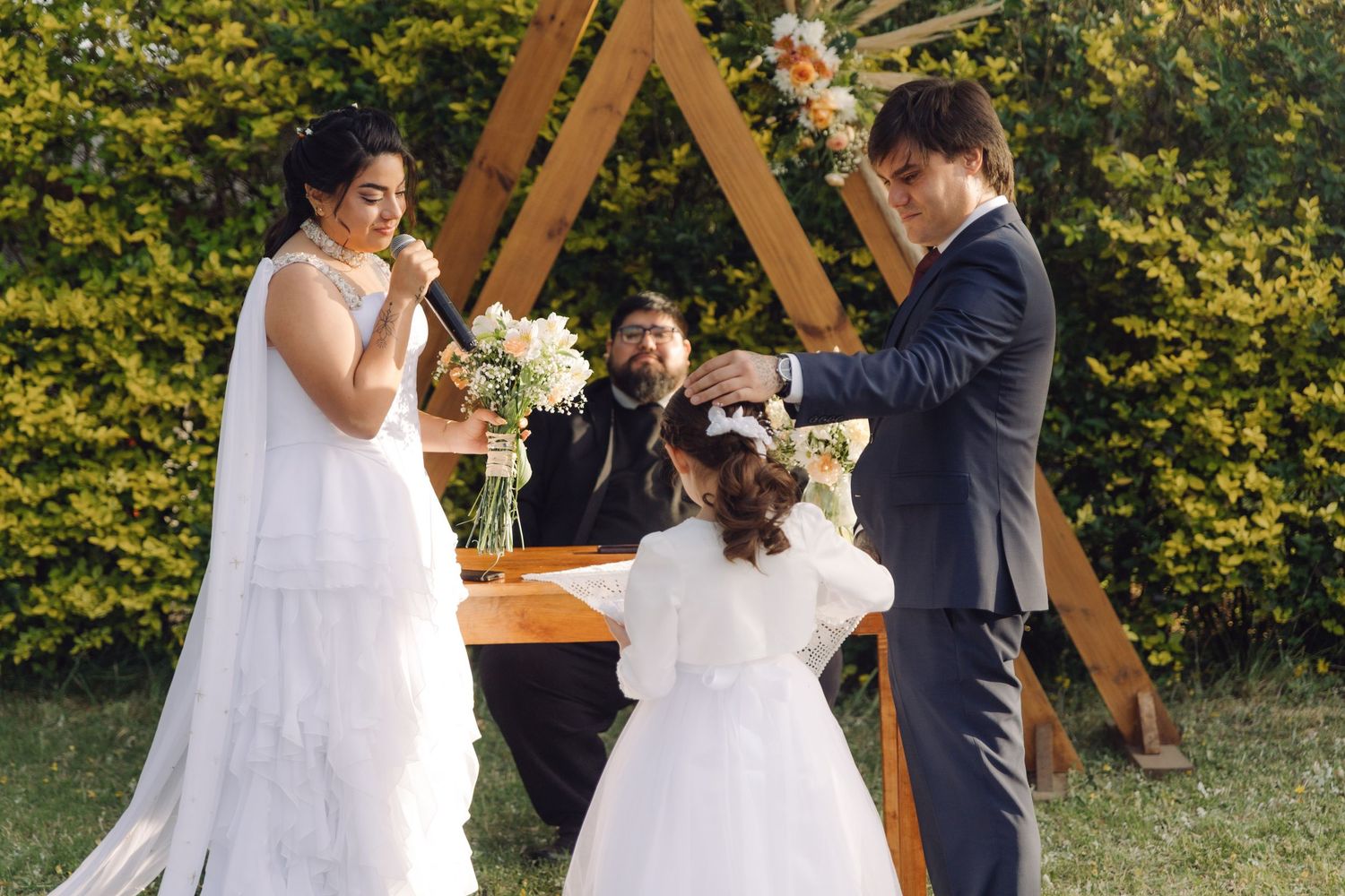 Groom places ring on brides hand during outdoor garden wedding ceremony with triangular wooden arch backdrop.
