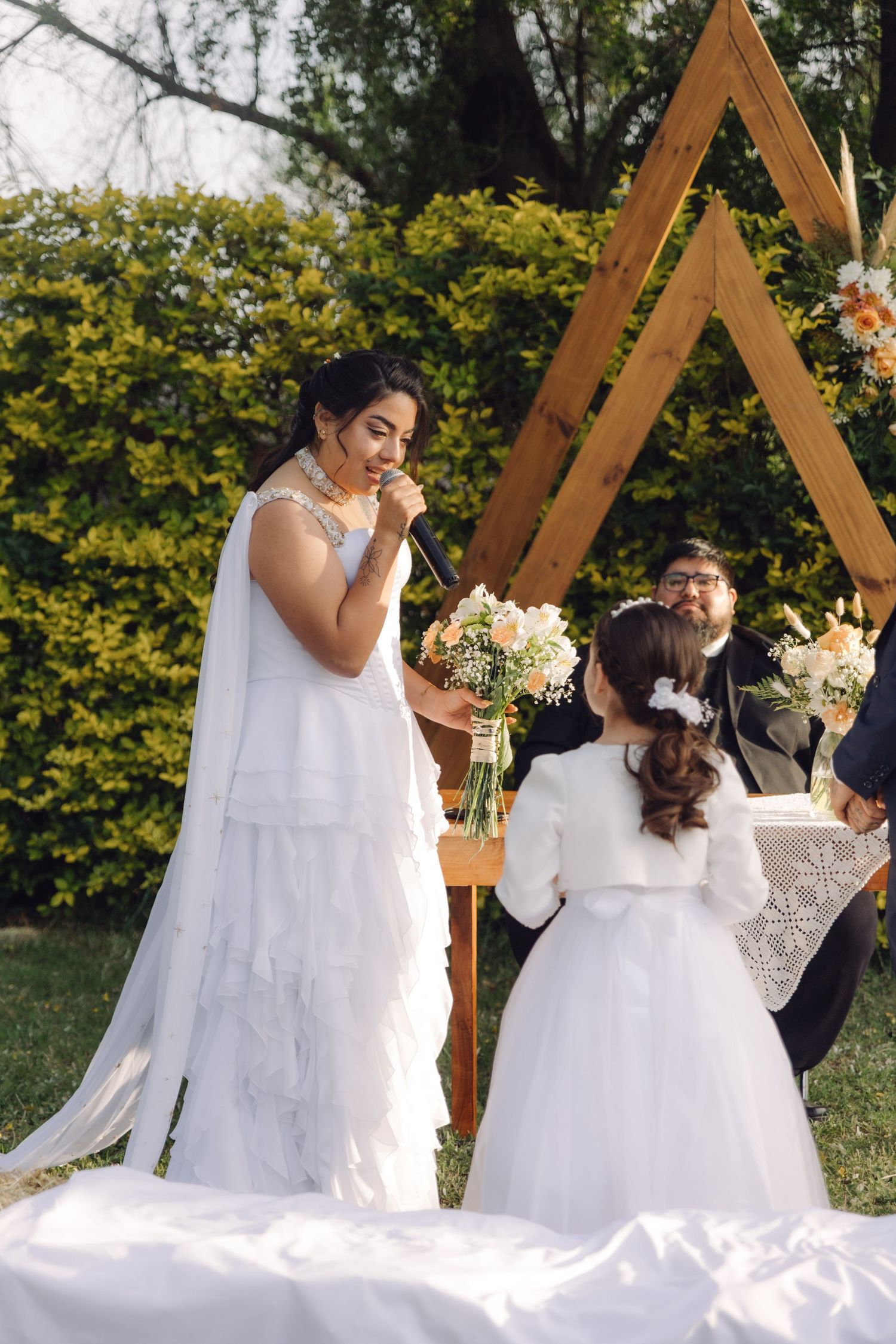 Bride smiles while speaking at outdoor wedding ceremony as flower girl watches near triangular wooden arch.