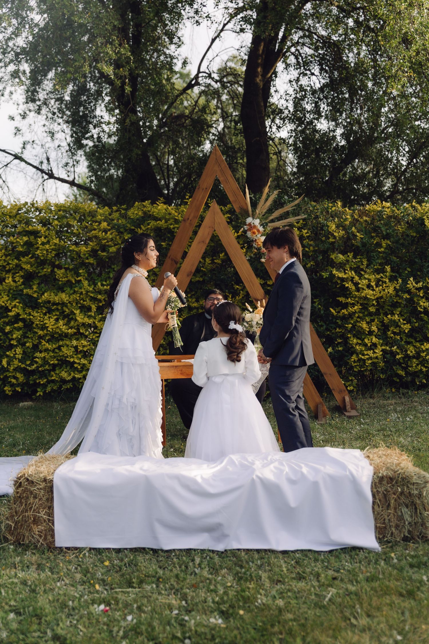Couple exchanges vows at an outdoor boho wedding ceremony with a triangular wooden arch and hay bale seating.