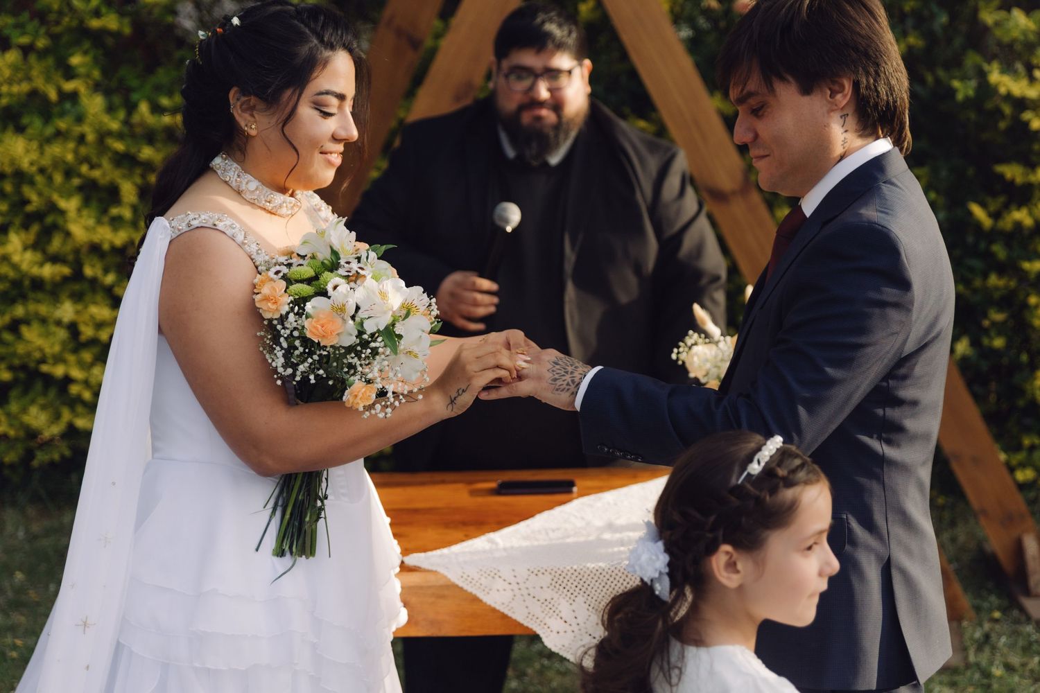 Bride and groom exchange rings at outdoor wedding ceremony with officiant and flower girl at wooden altar.