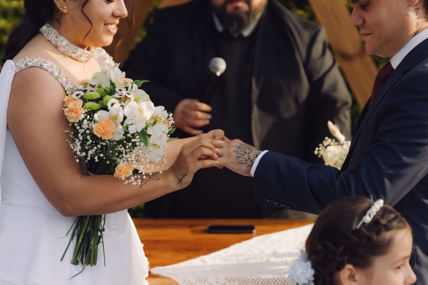 Bride and groom exchange rings during an outdoor wedding ceremony, bride holding a bouquet of white and peach roses.