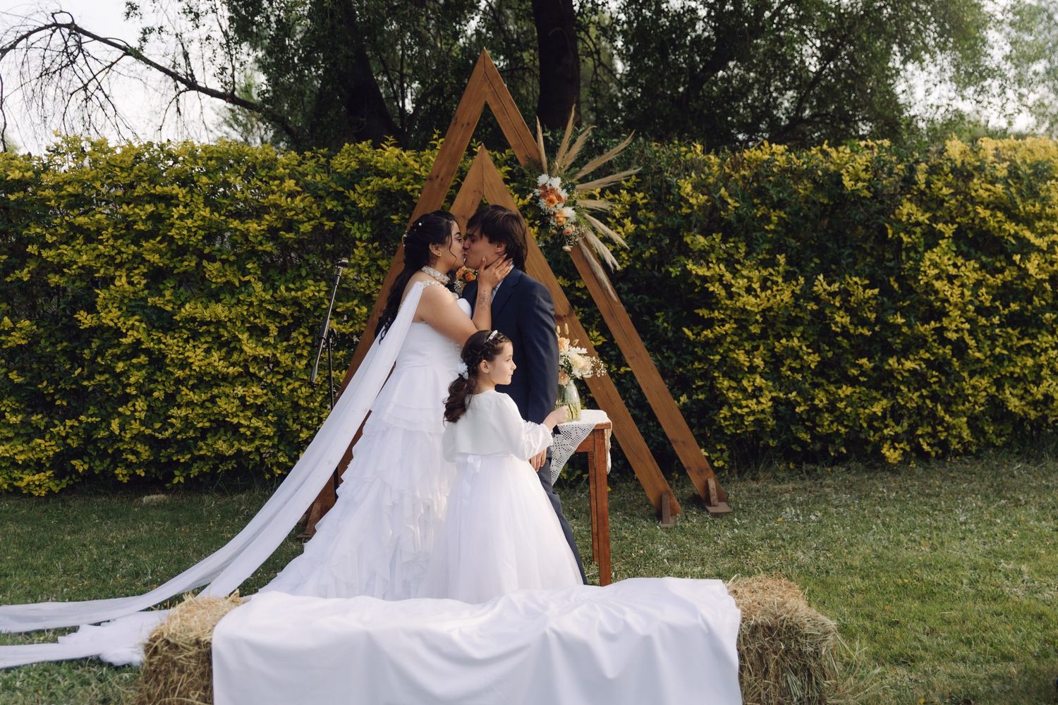 Bride and groom share their first kiss under a rustic triangular wooden arch at an outdoor garden wedding.