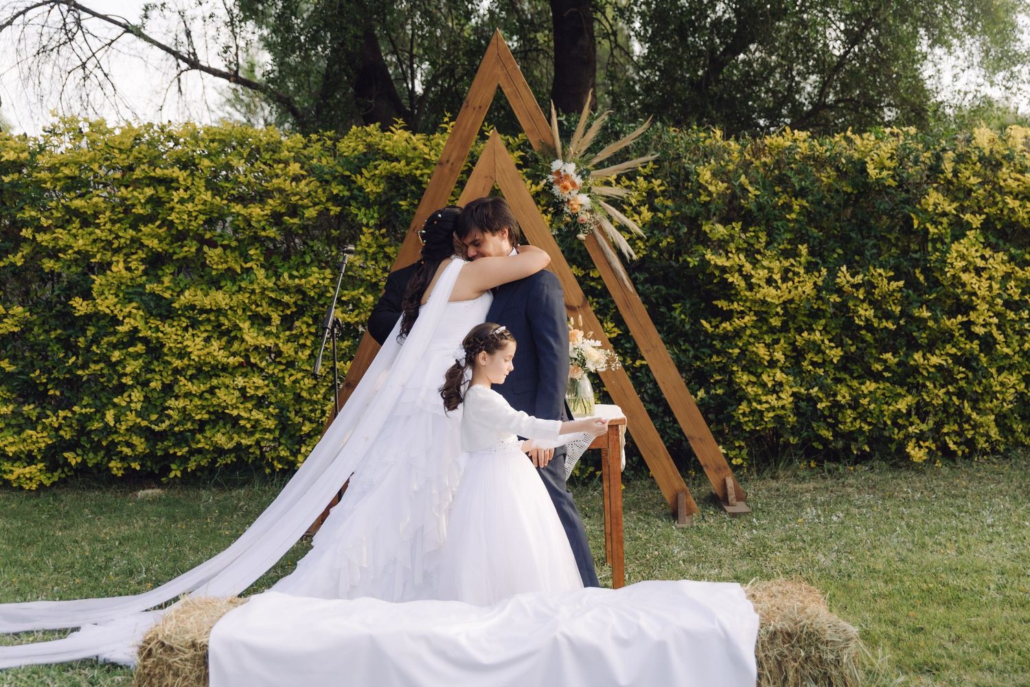 Bride and groom embrace at an outdoor boho wedding ceremony featuring a wooden triangle arch and lush green hedges.