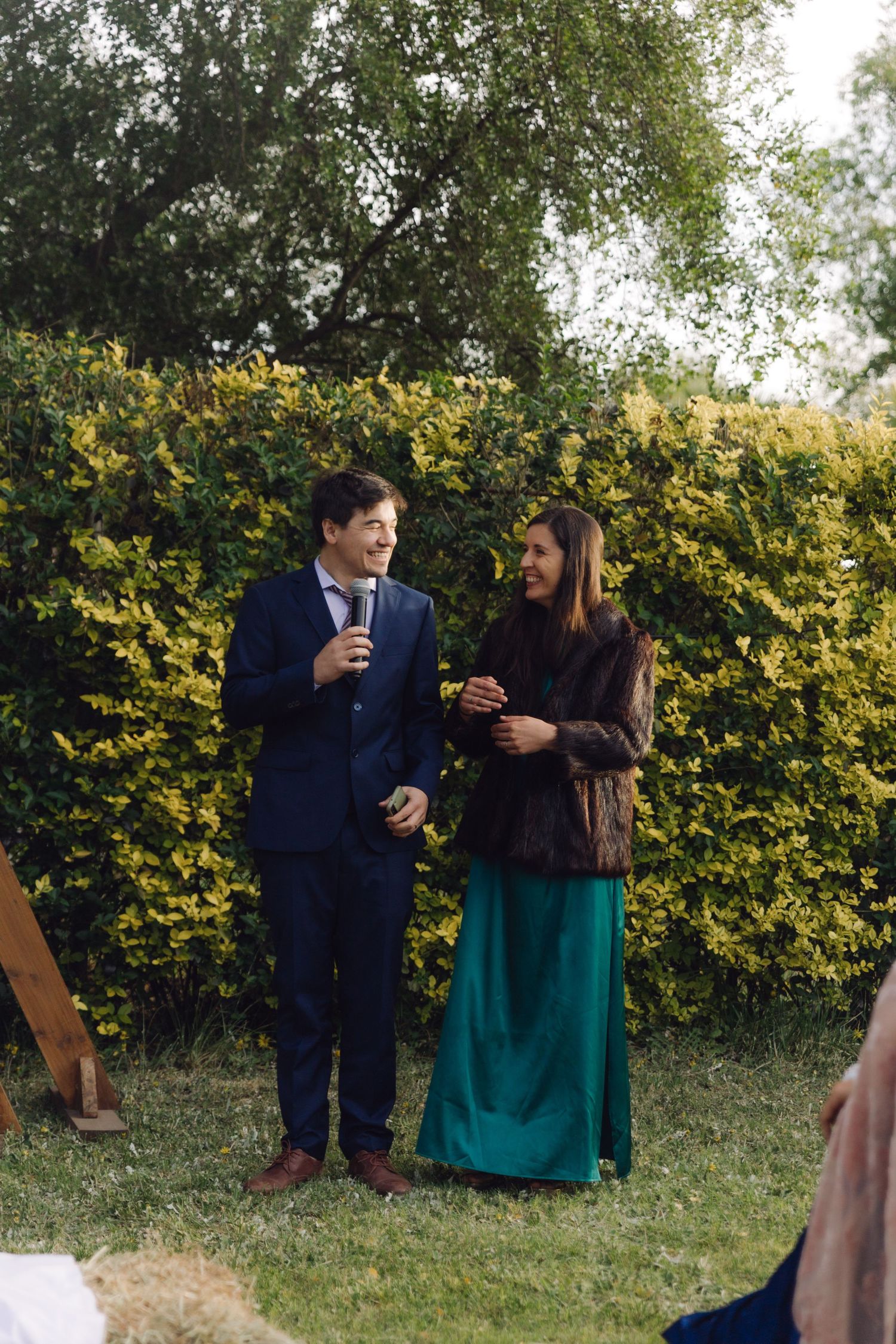 A young man in a navy suit and woman in a teal dress laugh together during an outdoor wedding ceremony.