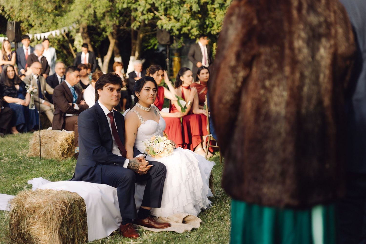Bride and groom sit on hay bales watching a speaker during their rustic outdoor wedding ceremony.