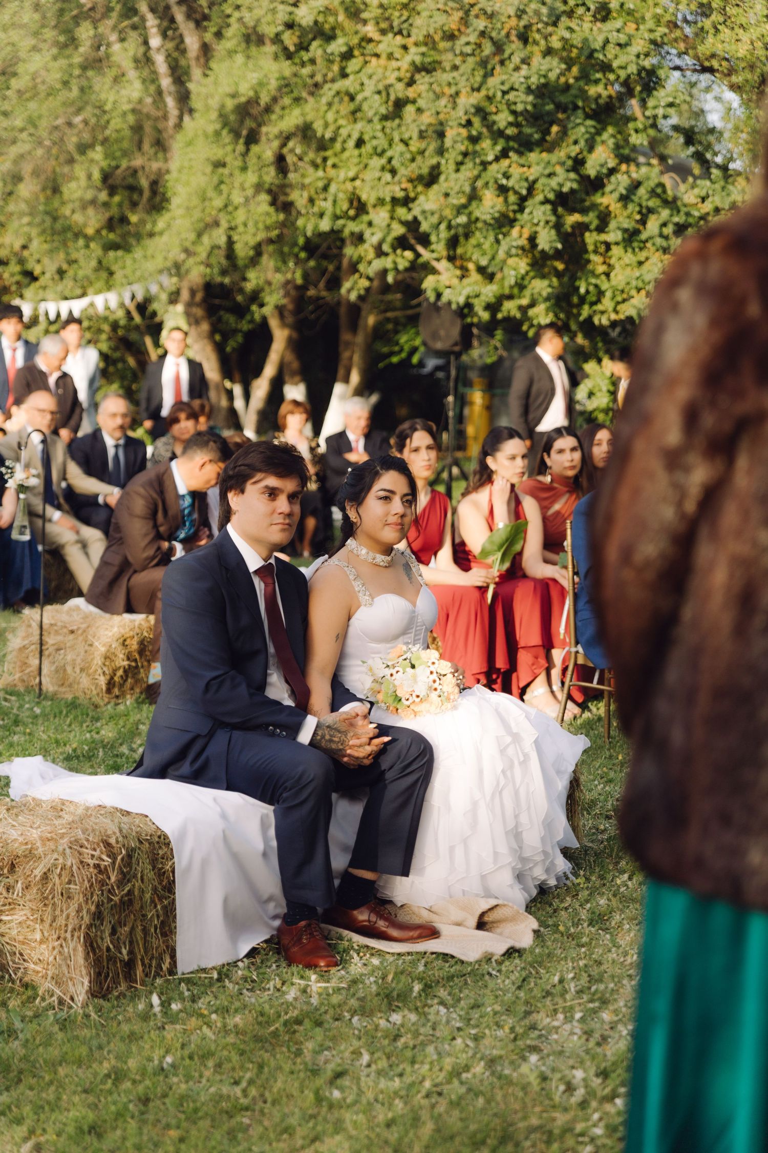 Bride in white gown and groom in navy suit sit on hay bales with wedding guests gathered in a garden.