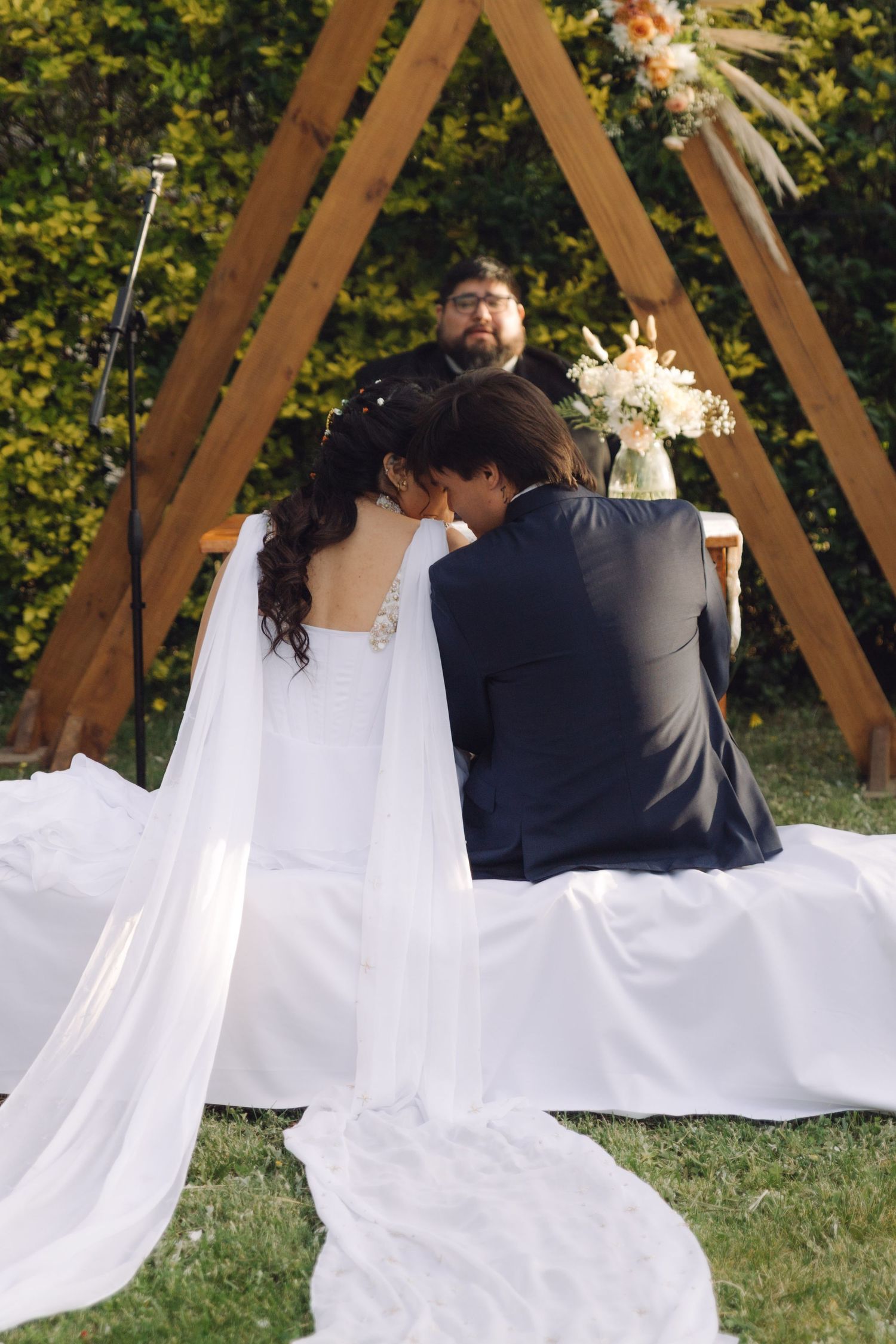 Bride and groom share an intimate moment seated at the altar before a wooden triangular arch outdoors.