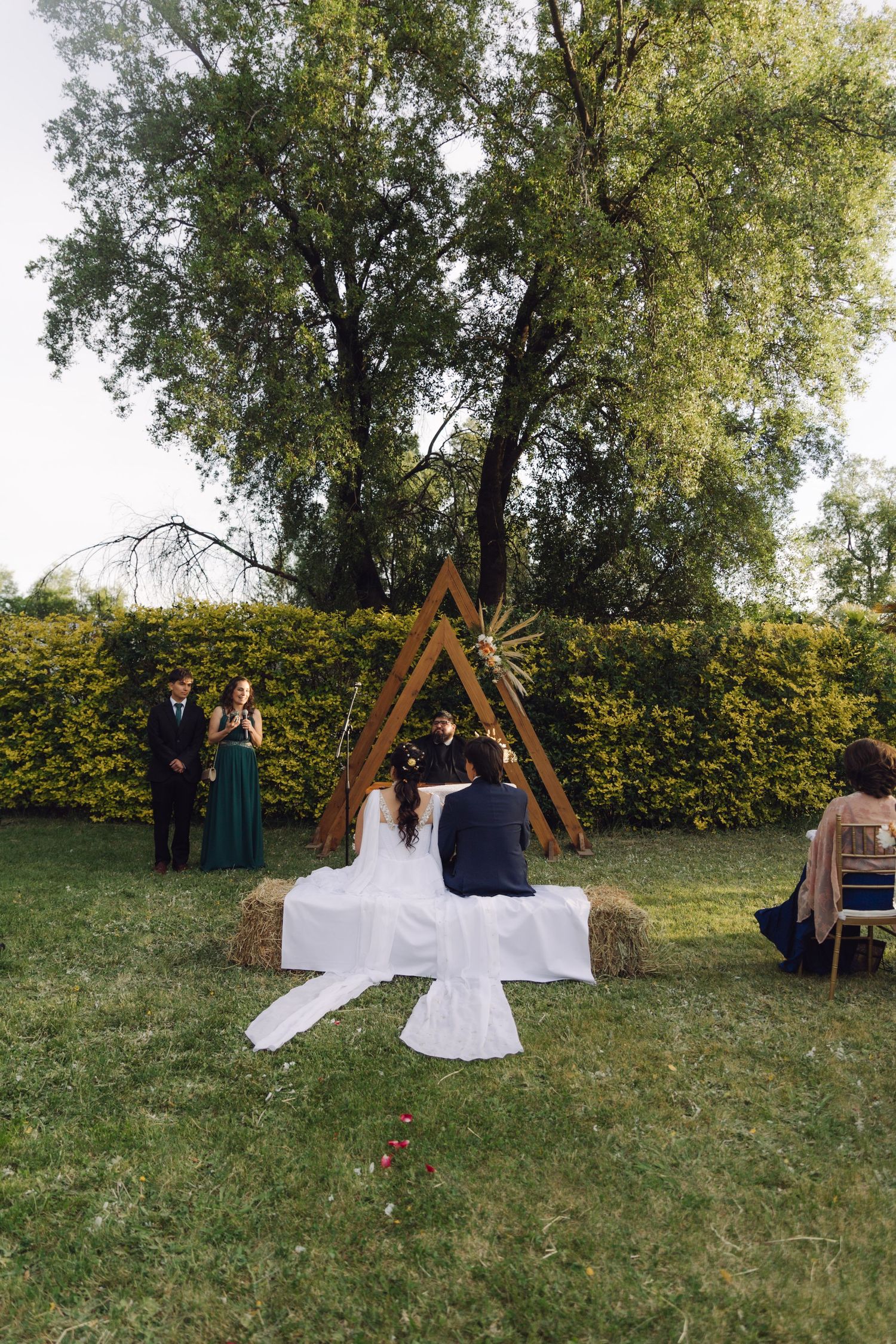 Bride and groom sit before a rustic triangular wooden arch during an intimate outdoor garden wedding ceremony.