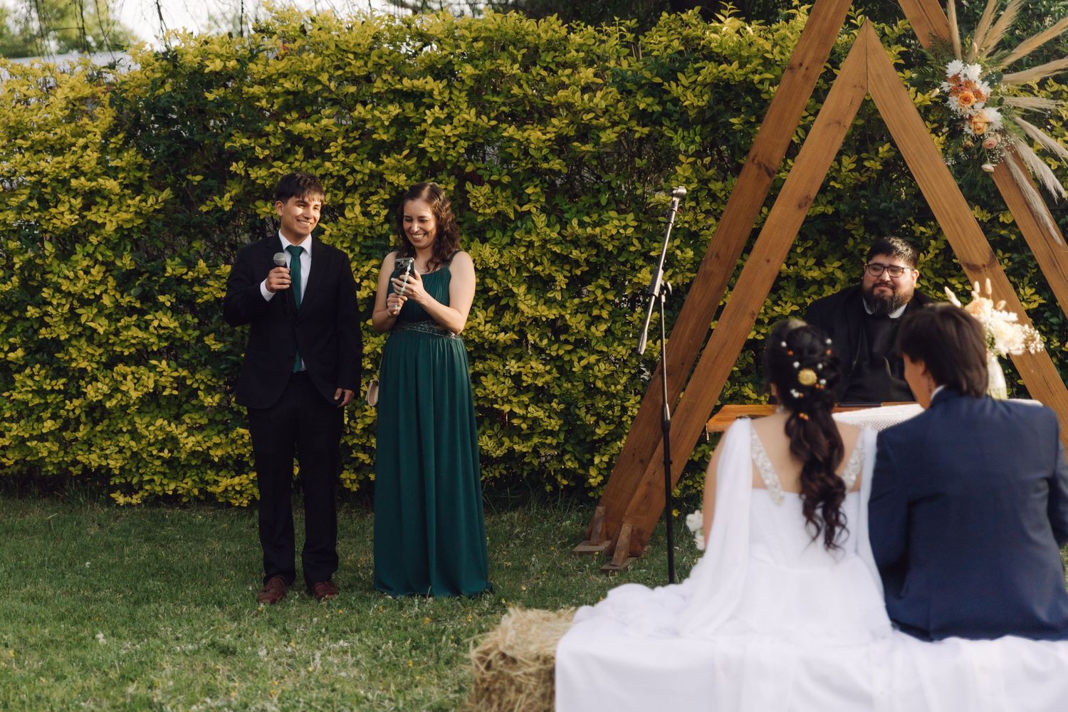 Two officiants speak at an outdoor bohemian wedding ceremony with a triangular wooden arch backdrop.