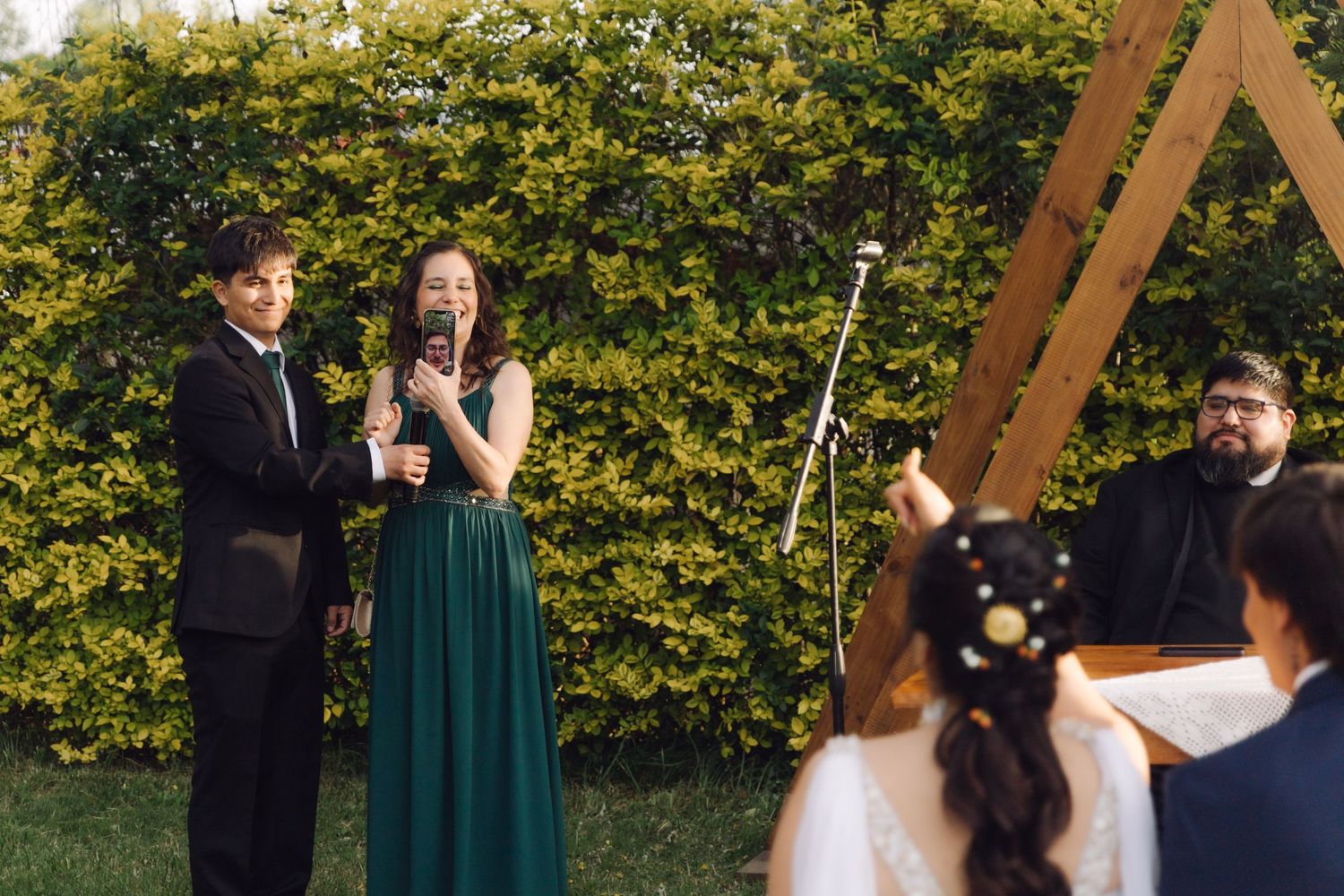 Two performers in green dresses sing into a microphone at an outdoor wedding ceremony near a wooden arch.