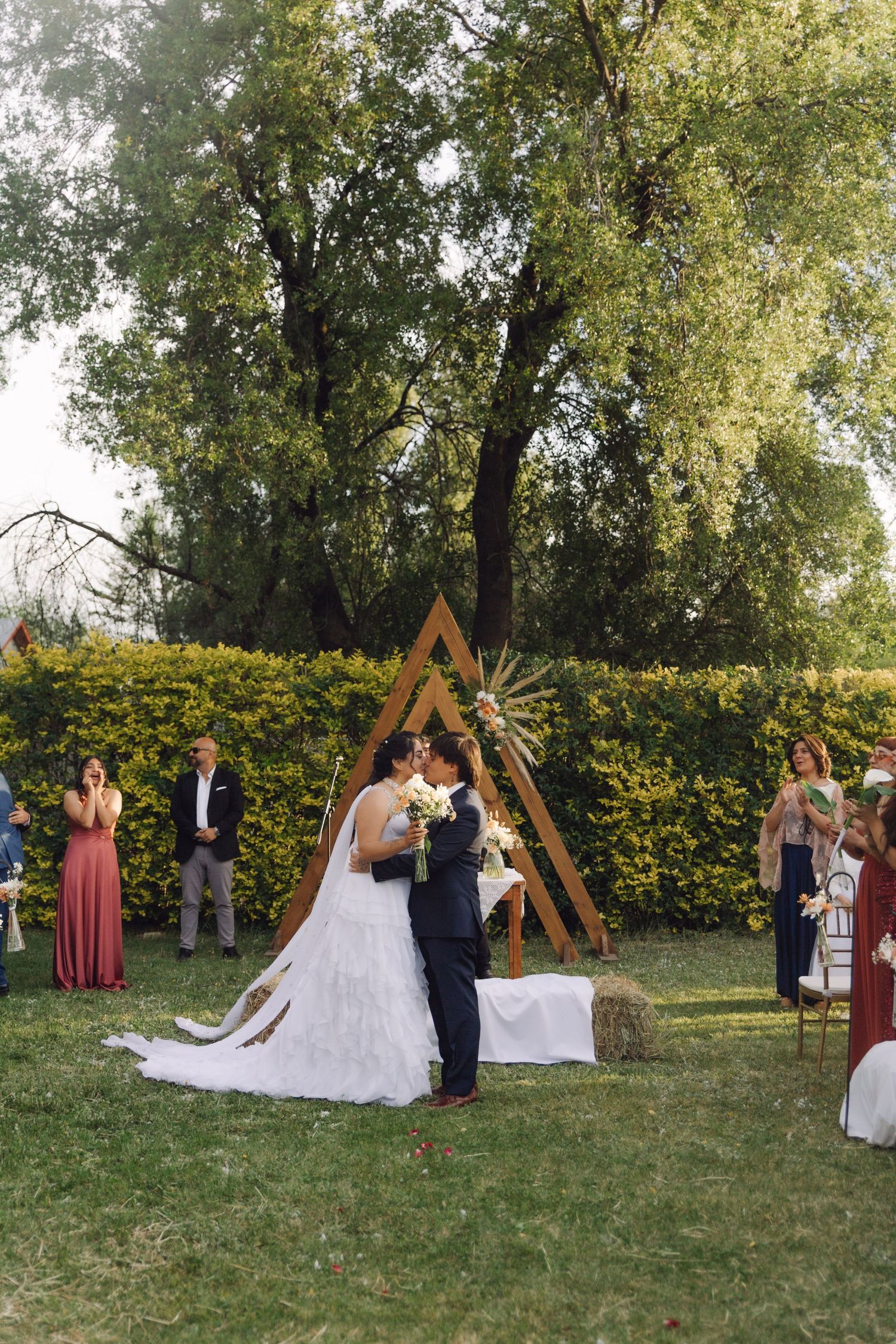 Bride and groom share first kiss at outdoor wedding under a triangular wooden arch with floral decor.
