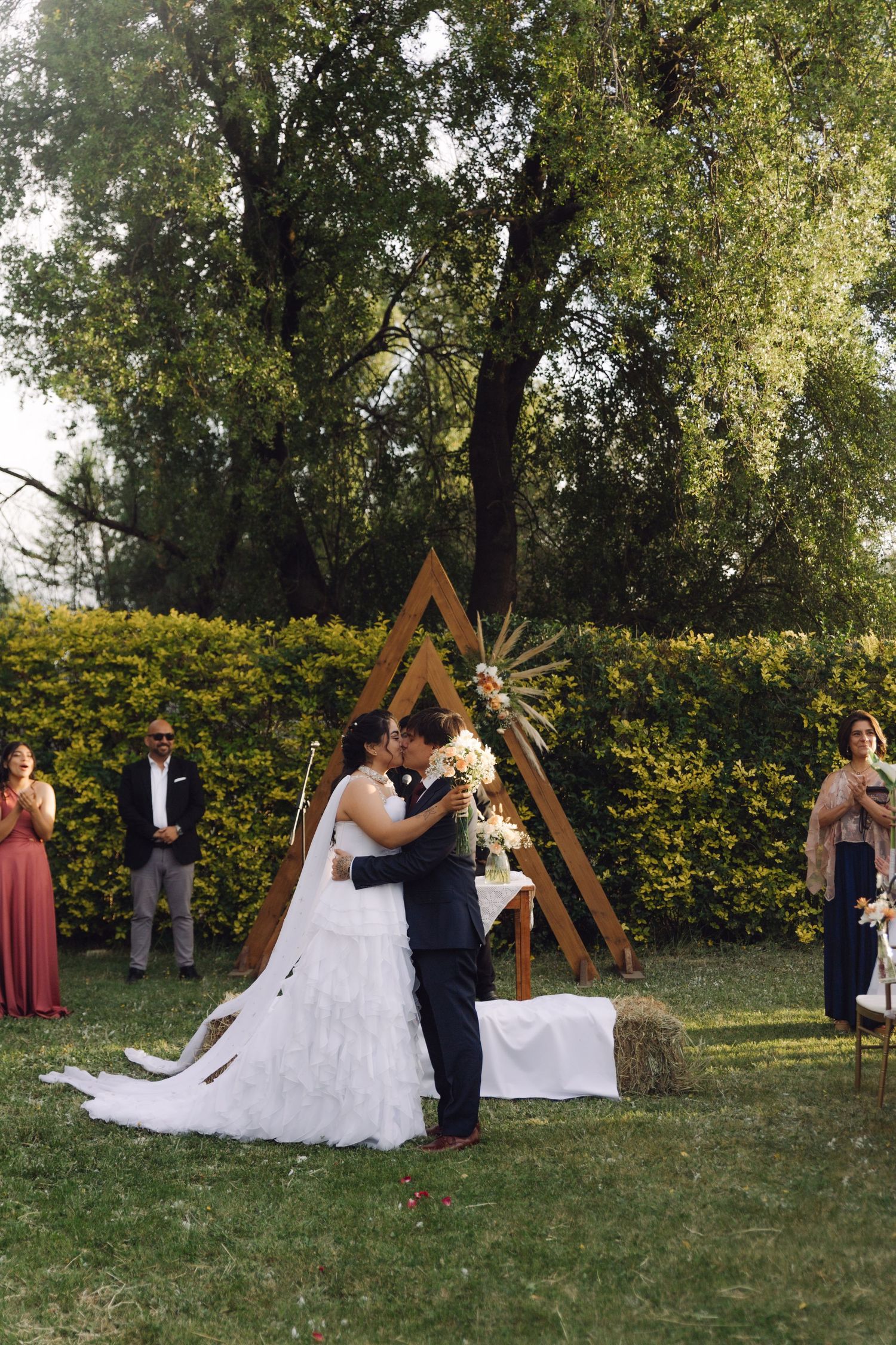 Bride and groom kiss at outdoor wedding ceremony with guests watching beside a wooden triangle arch.