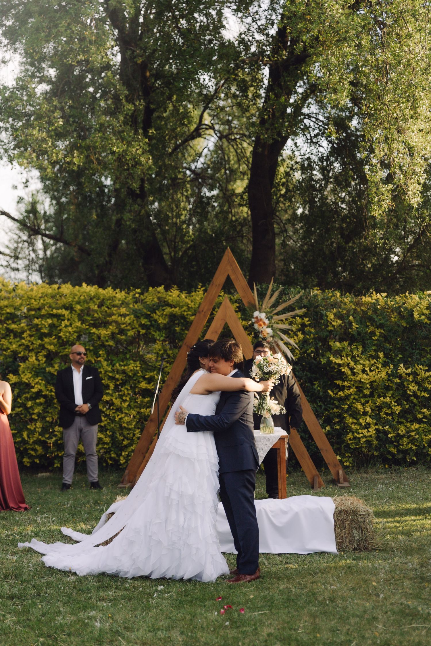 Newlyweds embrace at outdoor garden wedding ceremony in front of a rustic wooden triangle arch.